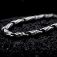 Load image into Gallery viewer, An oxidized Greek key chain necklace rests on dark volcanic rock, showing elongated engraved meander links and round connecting elements in a premium macro studio close-up.
