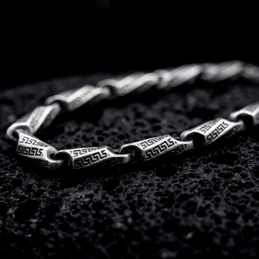 An oxidized Greek key chain necklace rests on dark volcanic rock, showing elongated engraved meander links and round connecting elements in a premium macro studio close-up.