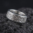 Load image into Gallery viewer, The polished silver Celtic interlace ring is displayed on textured black volcanic rock, highlighting the continuous knot pattern, smooth edges, and bold sterling silver band in a premium macro photograph.
