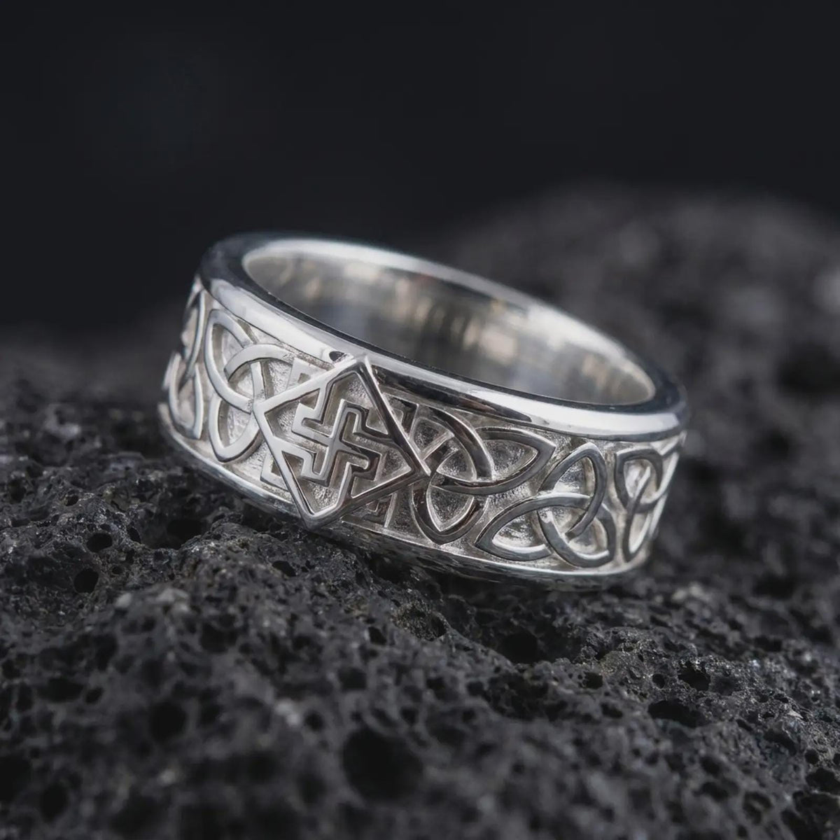 The polished silver Celtic interlace ring is displayed on textured black volcanic rock, highlighting the continuous knot pattern, smooth edges, and bold sterling silver band in a premium macro photograph.
