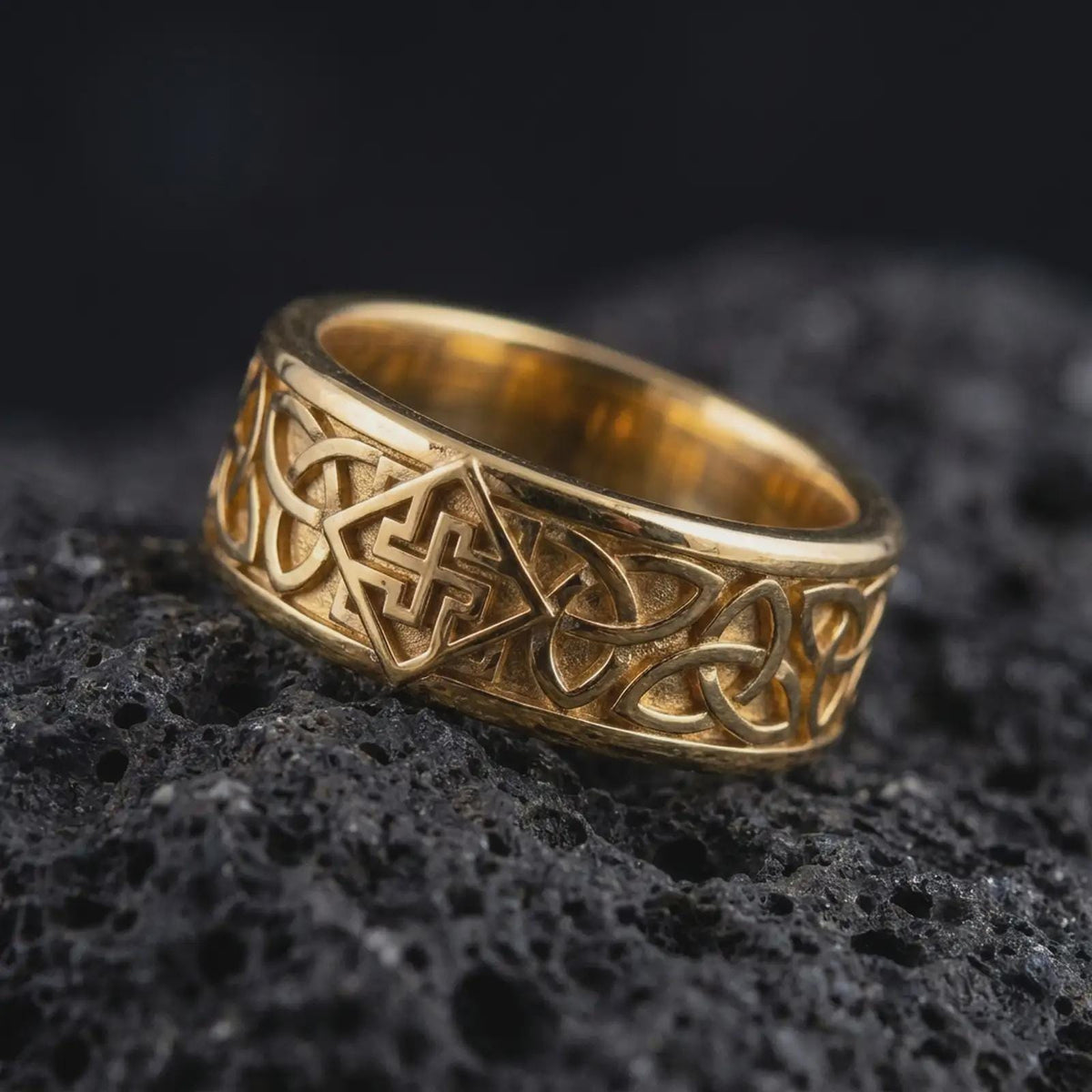 A gold plated Celtic knot band rests on rugged volcanic rock, featuring raised interwoven details and a symbolic continuous pattern across the wide ring in a luxury macro studio close-up.