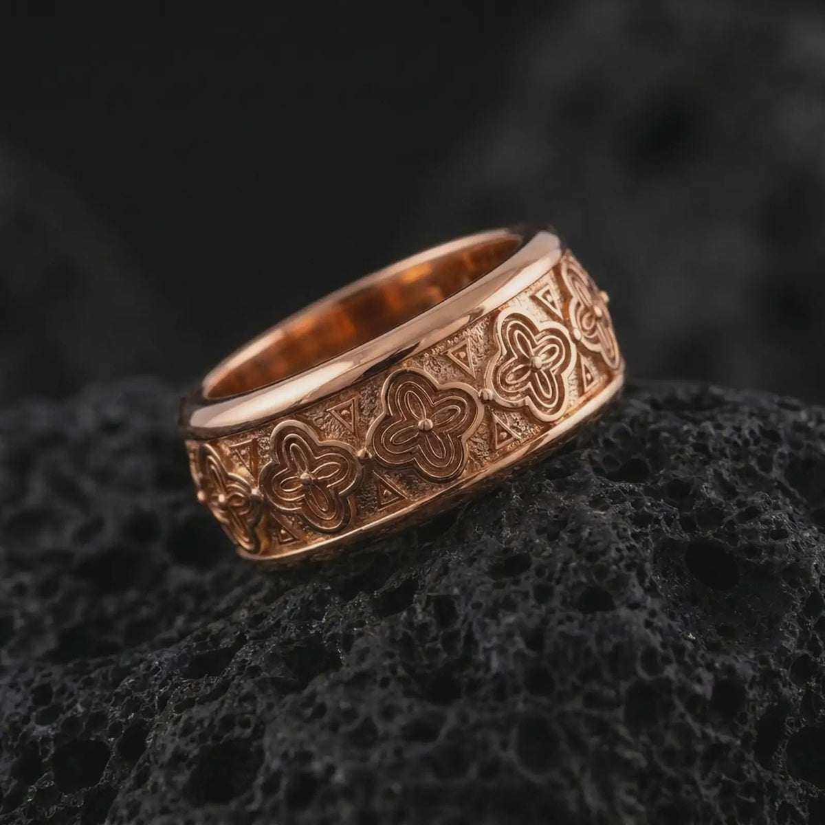 The rose gold plated floral wedding band is arranged on black volcanic rock, emphasizing its spinning ring construction, raised flower motifs, and refined unisex profile in a detailed macro image.