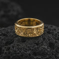 Load image into Gallery viewer, A gold plated spring flower ring rests on rugged volcanic rock, featuring raised blossoms, flowing ornamental lines, and a wide spinning band profile in a luxury macro studio close-up.
