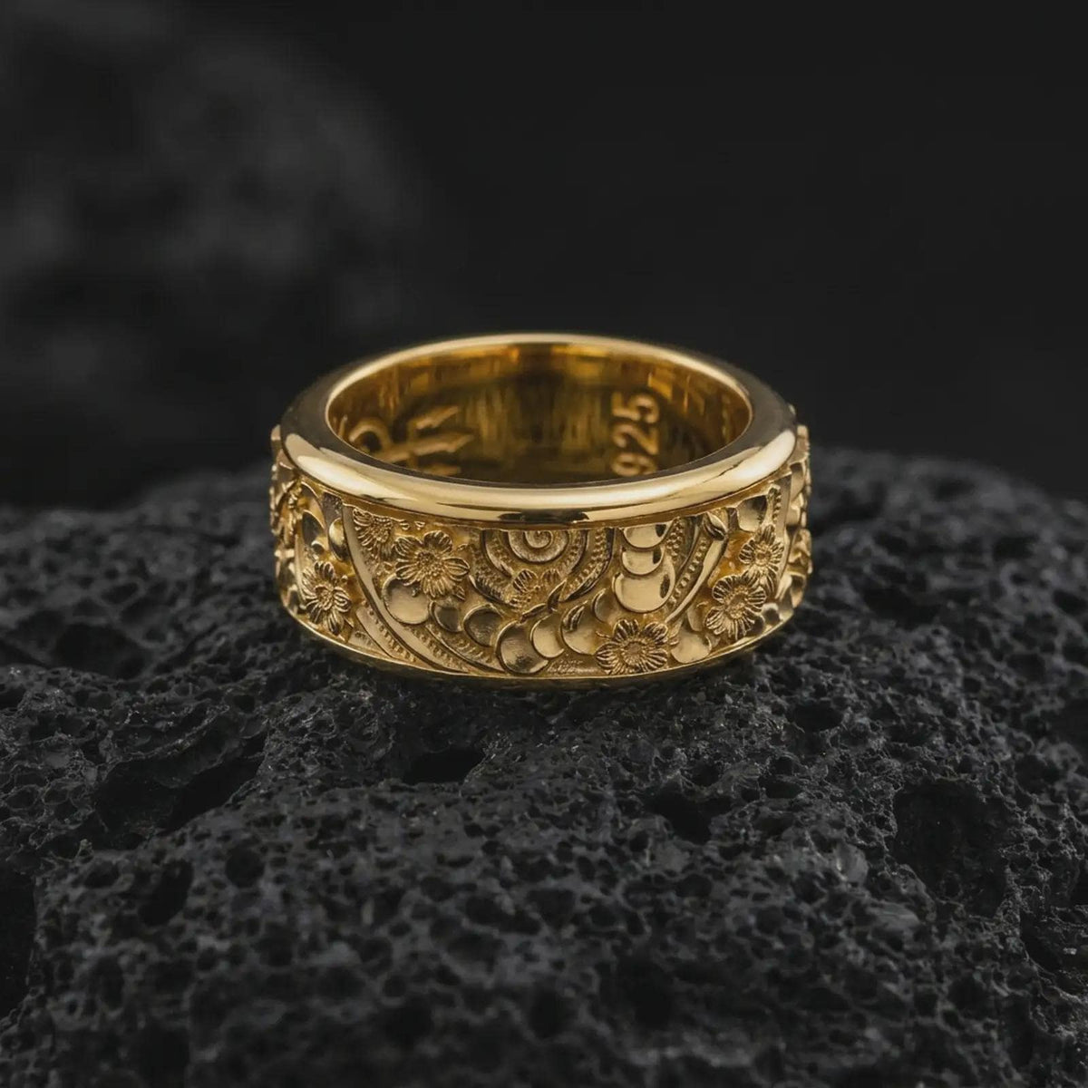 A gold plated spring flower ring rests on rugged volcanic rock, featuring raised blossoms, flowing ornamental lines, and a wide spinning band profile in a luxury macro studio close-up.