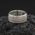 Load image into Gallery viewer, A polished silver Celtic knot wedding band is displayed on black volcanic rock, emphasizing the continuous knotwork, smooth rounded edges, and refined spinner band structure in a close-up image.

