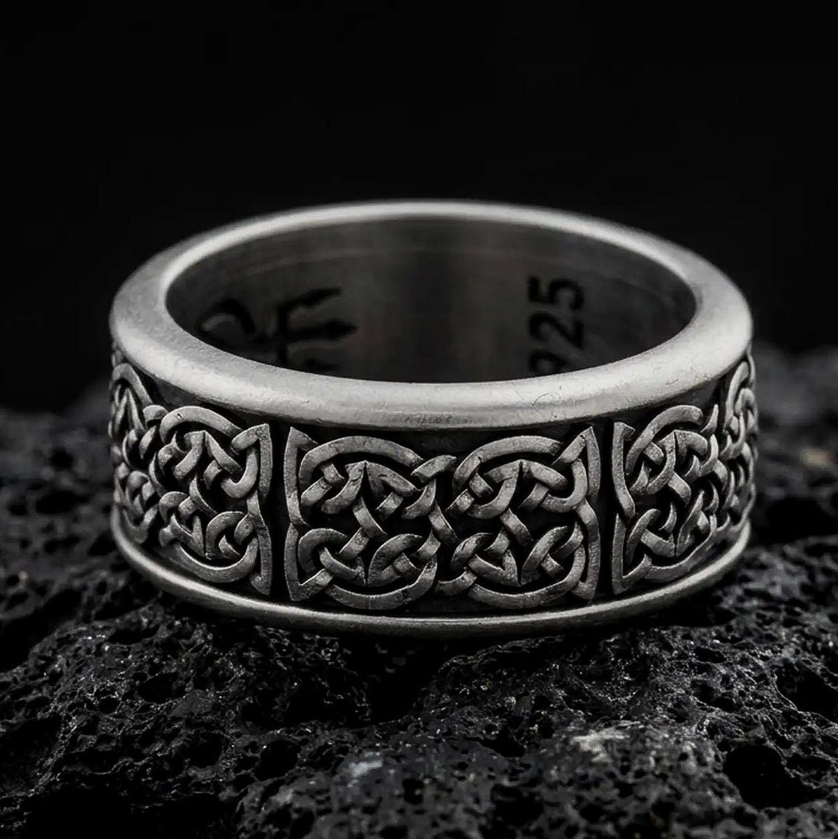 An oxidized sterling silver Celtic knot spinner ring rests on dark volcanic rock, highlighting the intricate interwoven Irish knot pattern and wide band design in a macro product photograph.