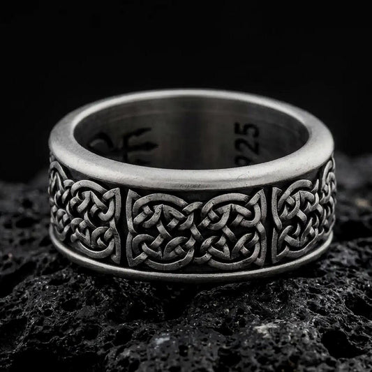 An oxidized sterling silver Celtic knot spinner ring rests on dark volcanic rock, highlighting the intricate interwoven Irish knot pattern and wide band design in a macro product photograph.