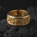 Load image into Gallery viewer, A gold plated floral wedding band rests on rugged volcanic rock, featuring raised flower motifs, flowing vine details, and a wide spinning ring profile in a luxury macro studio close-up.
