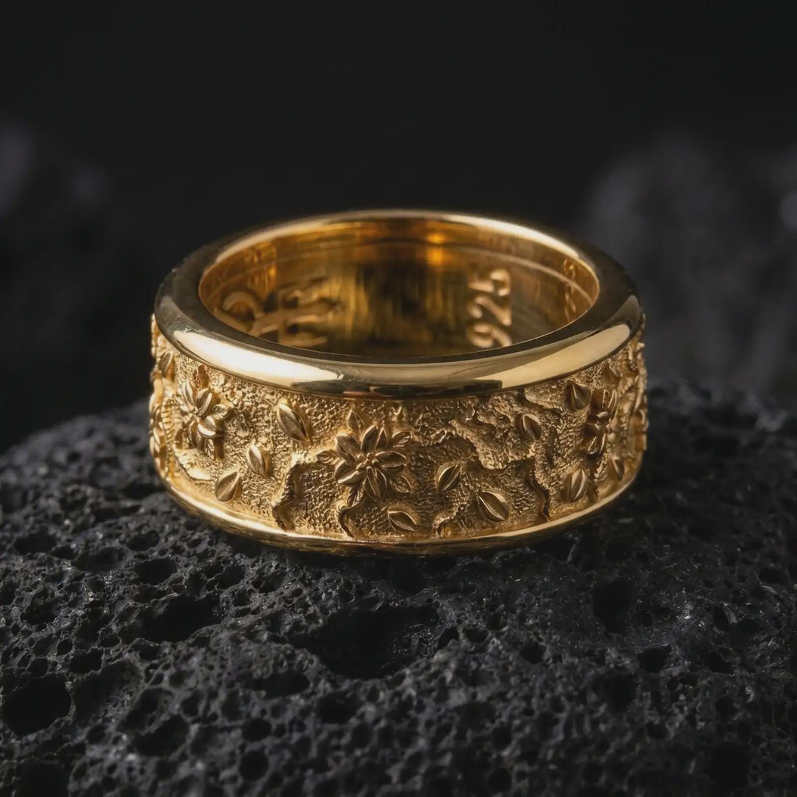 A gold plated floral wedding band rests on rugged volcanic rock, featuring raised flower motifs, flowing vine details, and a wide spinning ring profile in a luxury macro studio close-up.