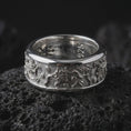 Load image into Gallery viewer, The polished silver flower spinner ring is displayed on textured black volcanic rock, highlighting the wide rounded band, botanical engravings, scattered leaves, and dimensional floral relief in a premium macro photograph.
