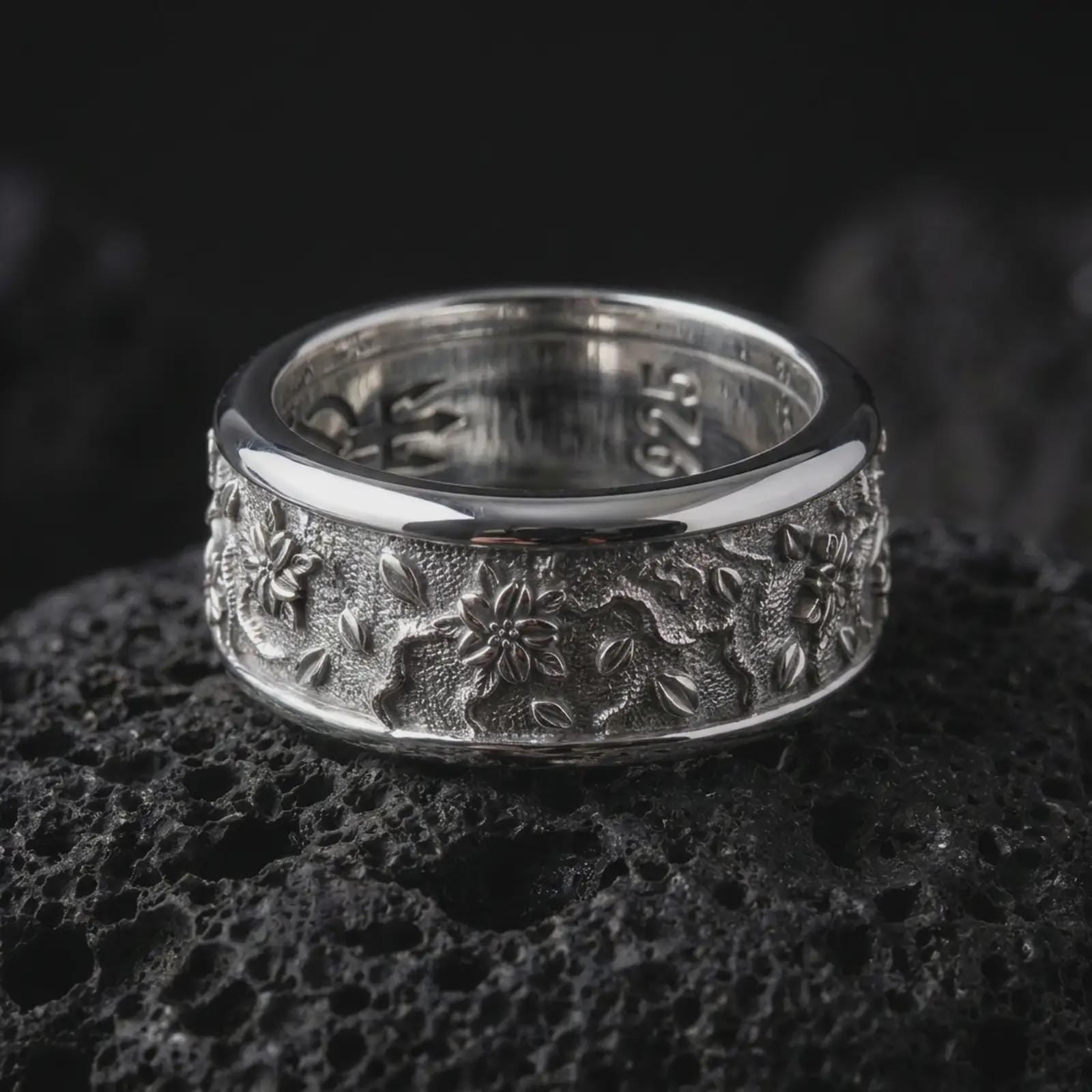 The polished silver flower spinner ring is displayed on textured black volcanic rock, highlighting the wide rounded band, botanical engravings, scattered leaves, and dimensional floral relief in a premium macro photograph.