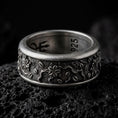 Load image into Gallery viewer, An oxidized sterling silver floral spinner ring rests on dark volcanic rock, showing raised blossoms, small leaf details, vine-like relief, and a textured recessed band background in a macro studio close-up.
