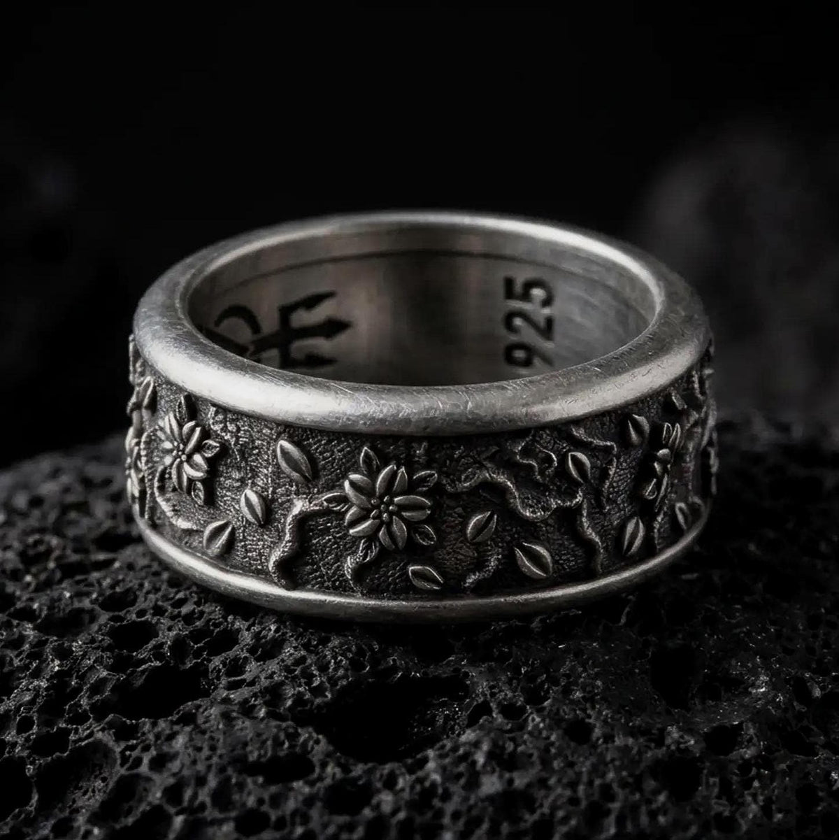 An oxidized sterling silver floral spinner ring rests on dark volcanic rock, showing raised blossoms, small leaf details, vine-like relief, and a textured recessed band background in a macro studio close-up.