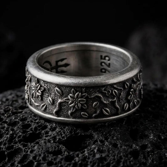 An oxidized sterling silver floral spinner ring rests on dark volcanic rock, showing raised blossoms, small leaf details, vine-like relief, and a textured recessed band background in a macro studio close-up.