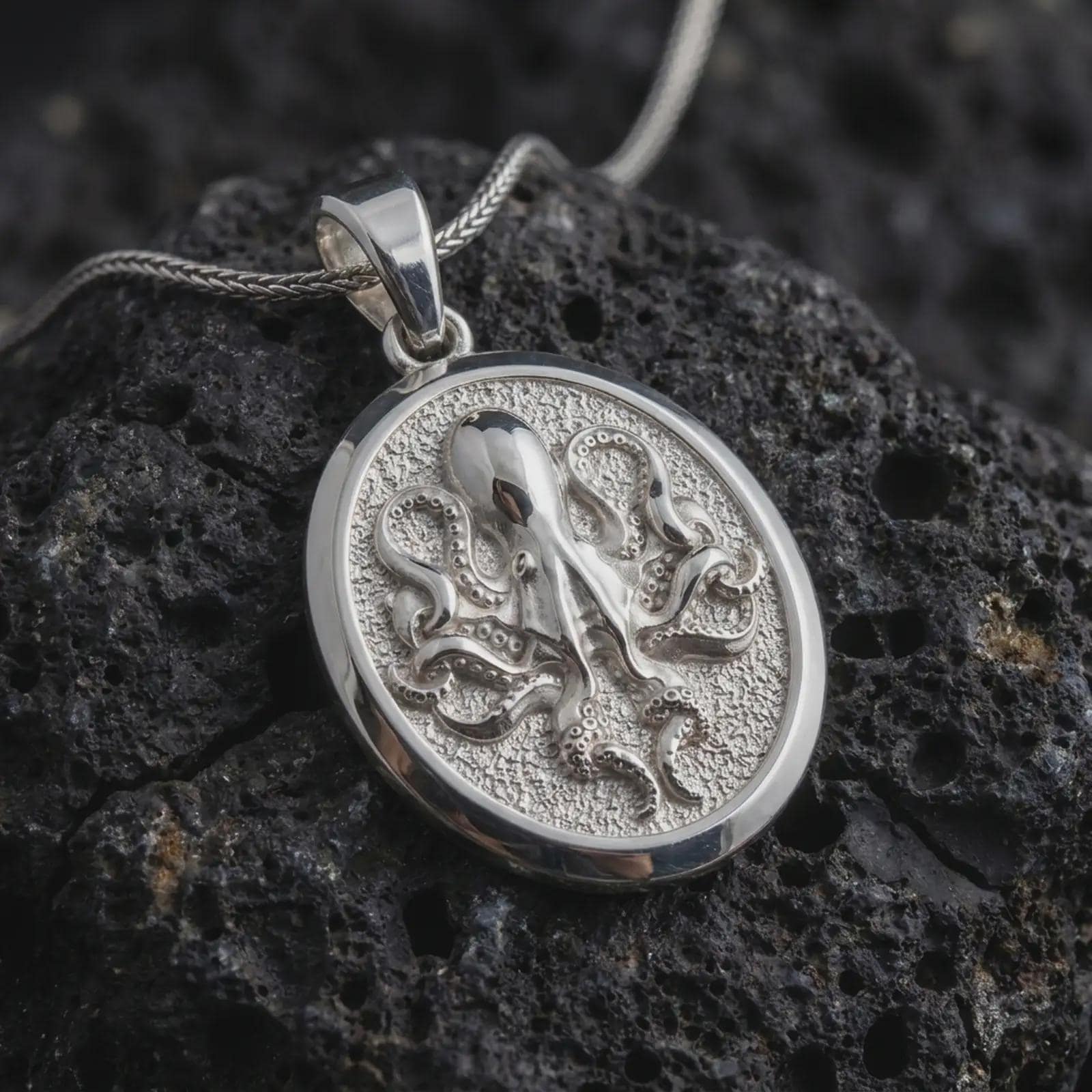 The polished silver octopus necklace is displayed on textured black volcanic rock, highlighting the dimensional sea creature, flowing tentacles, smooth circular frame, and detailed marine relief in a premium macro photograph.