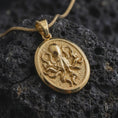 Load image into Gallery viewer, A gold plated octopus pendant lies on rugged volcanic rock, featuring a raised kraken-inspired body, curling tentacles, and a dark textured ocean-style background in a luxury macro studio close-up.
