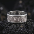 Load image into Gallery viewer, The polished silver Aztec pattern ring is displayed on textured black volcanic rock, highlighting the repeating engraved motifs, smooth rounded edges, and spinning band profile in a premium macro photograph.
