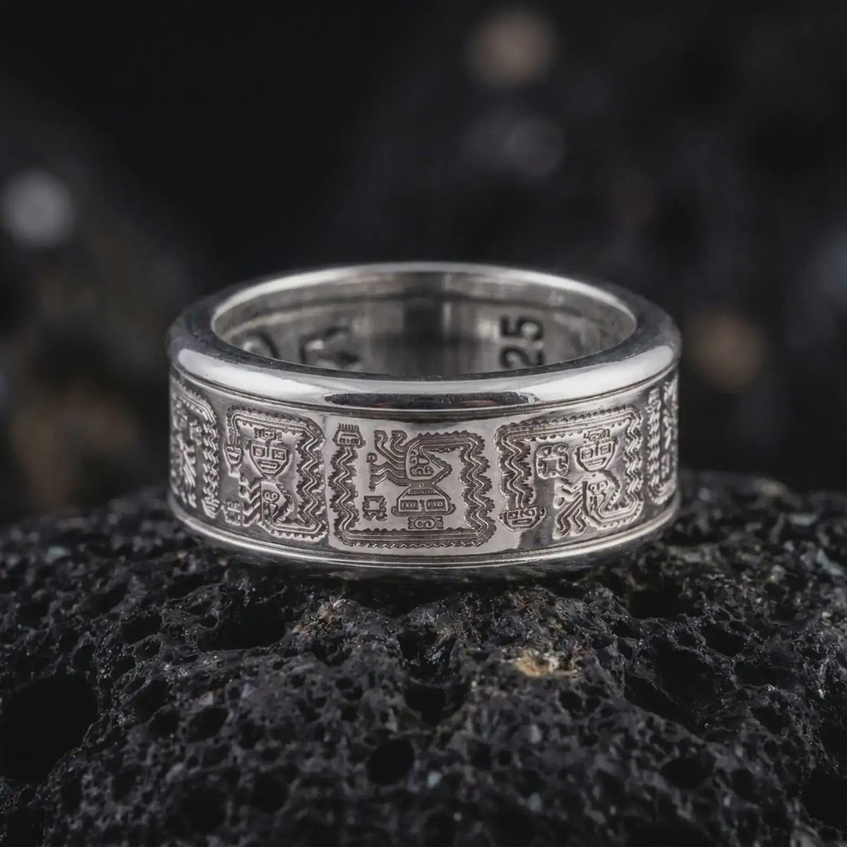 The polished silver Aztec pattern ring is displayed on textured black volcanic rock, highlighting the repeating engraved motifs, smooth rounded edges, and spinning band profile in a premium macro photograph.