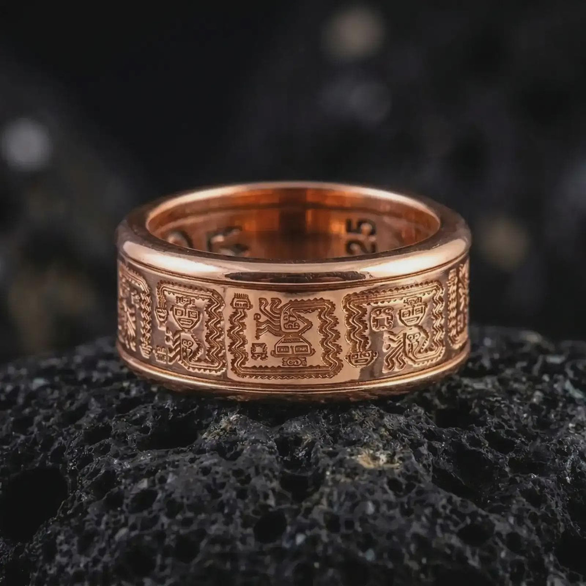The rose gold plated Aztec pattern ring is arranged on black volcanic rock, emphasizing its tribal-inspired engravings, textured recessed background, and refined unisex spinner band design in a detailed macro image.