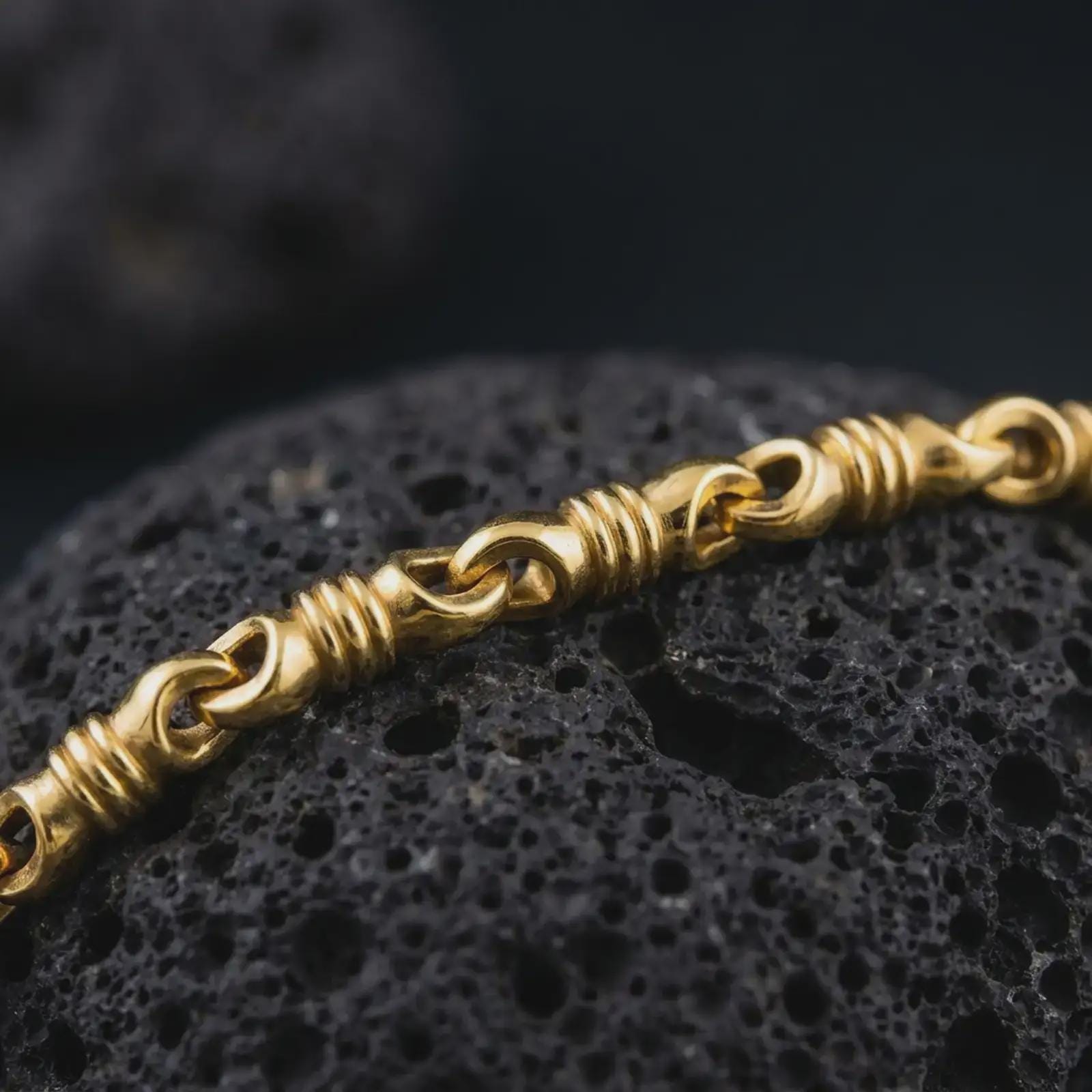 A gold plated ridged barrel necklace lies across rugged volcanic rock, featuring repeating cylinder links, rounded connectors, and a bold masculine chain profile in a luxury macro studio close-up.
