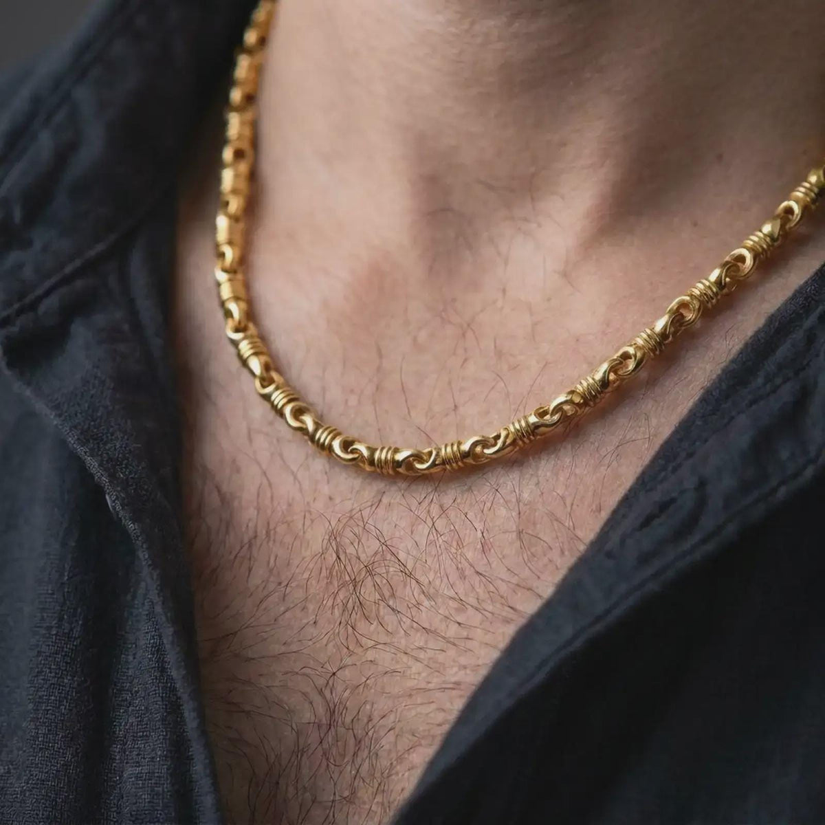 A gold plated barrel chain necklace is worn on a man’s neck with dark clothing, highlighting the sculpted ridges, rounded link pattern, and strong statement style in a masculine close-up.