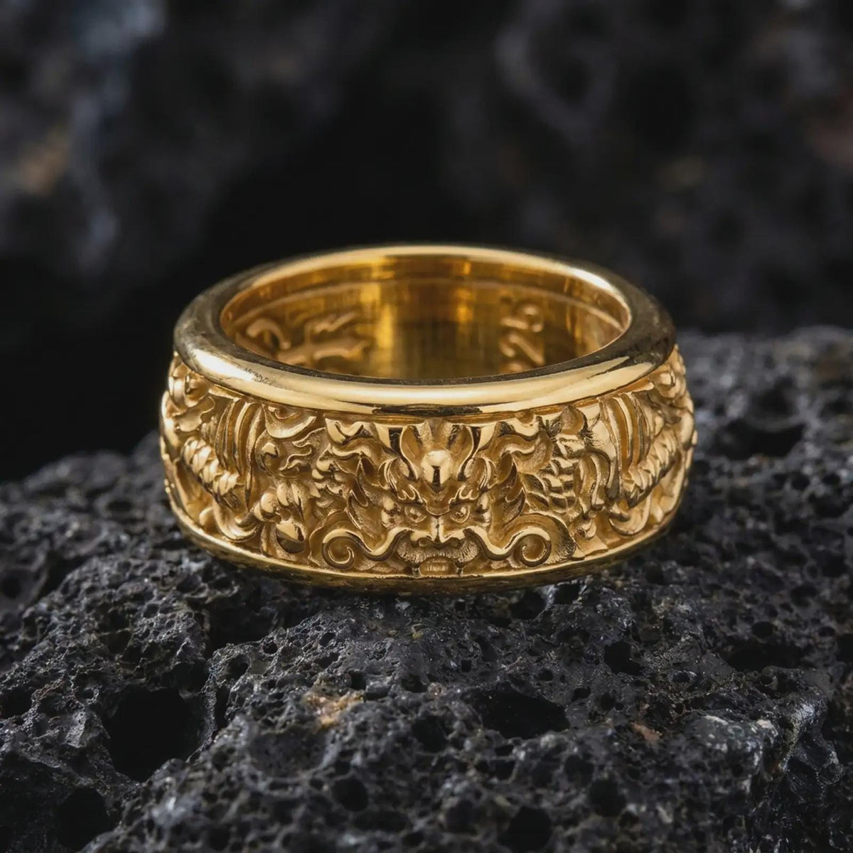A gold plated Asian dragon spinner ring rests on rugged volcanic rock, featuring ornate dragon motifs, symbolic craftsmanship, and an engravable wide band in a luxury macro studio close-up.