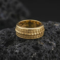 Load image into Gallery viewer, A gold plated lizard scale spinner ring sits on textured volcanic rock, showcasing the raised reptile motif, bold rotating band, and striking statement jewelry design in a luxury macro studio image.
