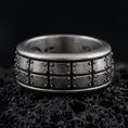 Load image into Gallery viewer, An oxidized sterling silver plate pattern spinner ring rests on dark volcanic rock, highlighting the geometric panel design, rotating band structure, and rugged industrial texture in a dramatic macro close-up.
