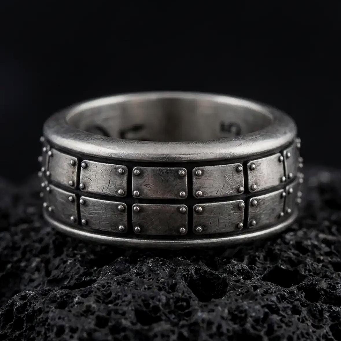 An oxidized sterling silver plate pattern spinner ring rests on dark volcanic rock, highlighting the geometric panel design, rotating band structure, and rugged industrial texture in a dramatic macro close-up.