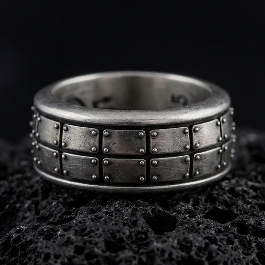 An oxidized sterling silver plate pattern spinner ring rests on dark volcanic rock, highlighting the geometric panel design, rotating band structure, and rugged industrial texture in a dramatic macro close-up.