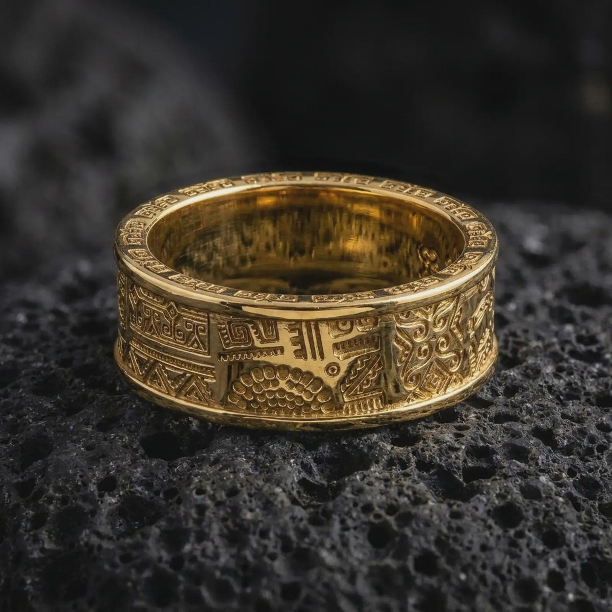 A gold plated Aztec spinner ring rests on rugged volcanic rock, featuring symbolic panel engravings, stepped linework, and a bold unisex wedding band profile in a luxury macro studio close-up.