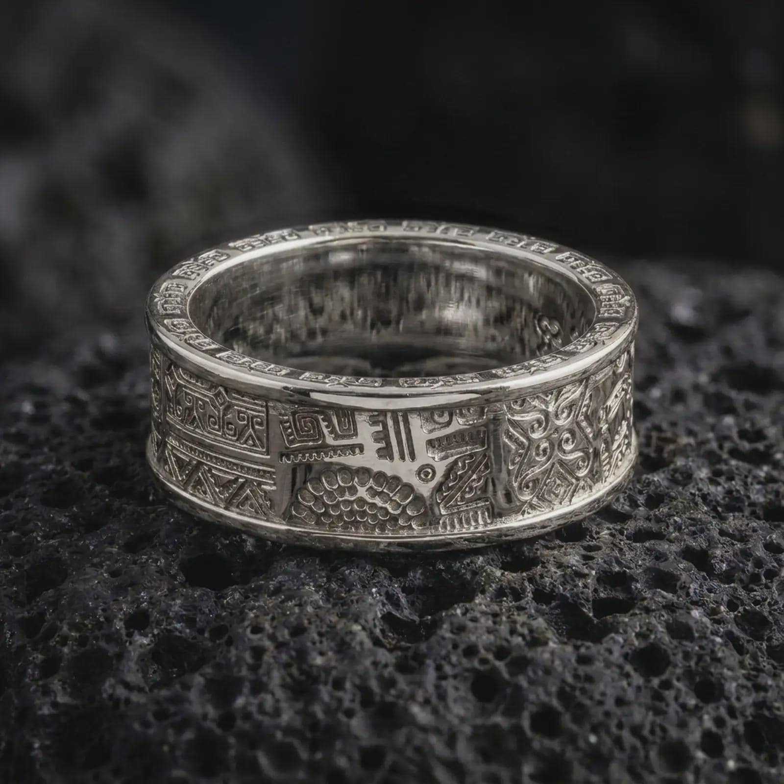 The polished silver Aztec pattern ring is displayed on textured black volcanic rock, highlighting the wide spinning band, engraved geometric details, and smooth rounded edges in a premium macro photograph.
