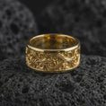 Load image into Gallery viewer, A gold plated vine scroll ring rests on rugged volcanic rock, featuring raised botanical curves, dark engraved depth, and a wide spinning band profile in a luxury macro studio close-up.
