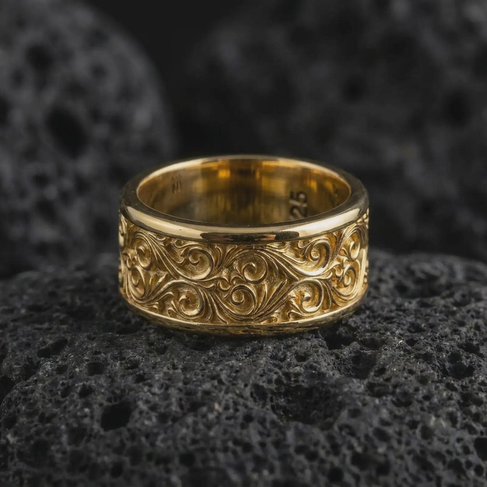 A gold plated vine scroll ring rests on rugged volcanic rock, featuring raised botanical curves, dark engraved depth, and a wide spinning band profile in a luxury macro studio close-up.