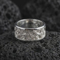 Load image into Gallery viewer, The polished silver ornate spinner ring is displayed on textured black volcanic rock, highlighting the flowing scroll pattern, smooth rounded edges, and wide sterling silver band in a premium macro photograph.
