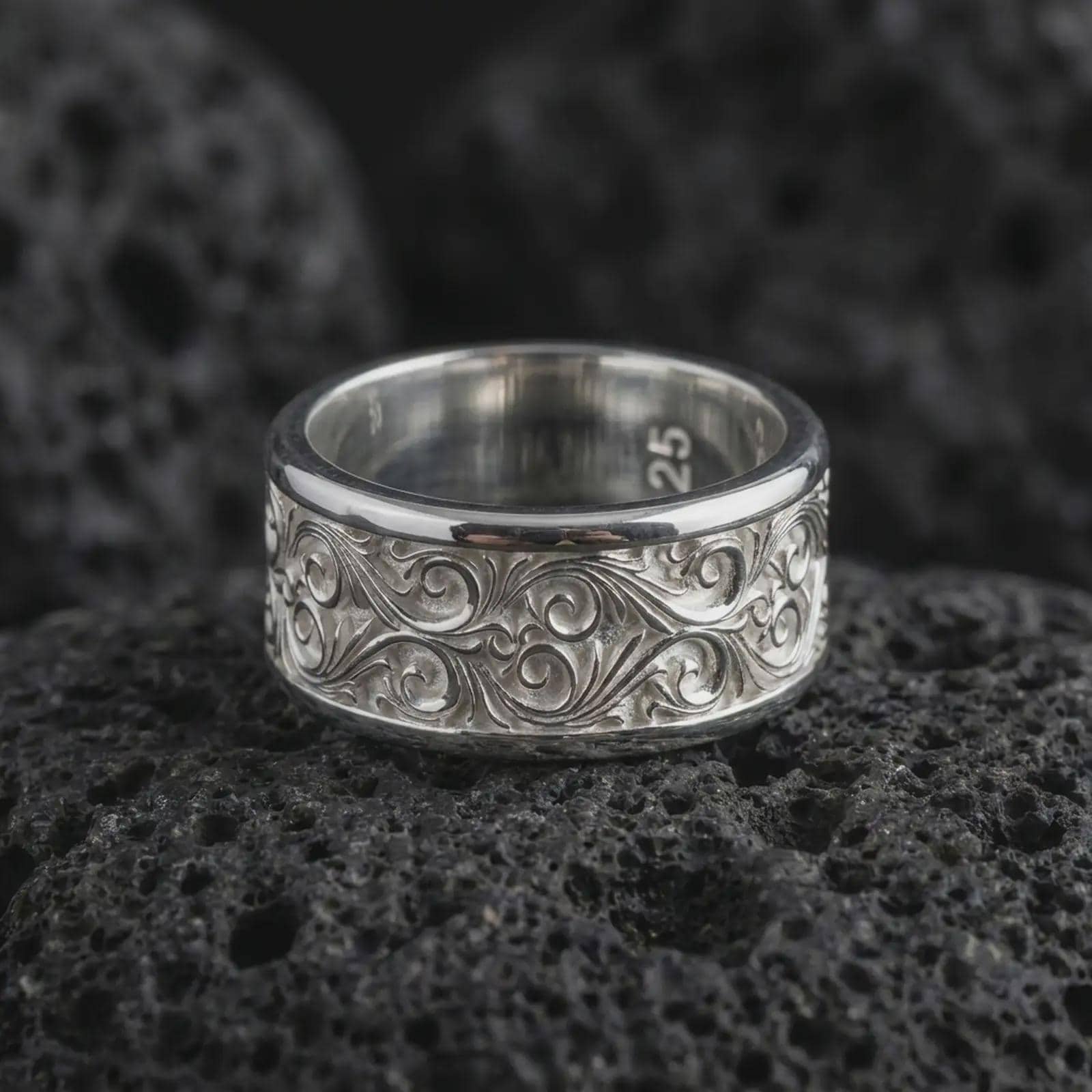 The polished silver ornate spinner ring is displayed on textured black volcanic rock, highlighting the flowing scroll pattern, smooth rounded edges, and wide sterling silver band in a premium macro photograph.