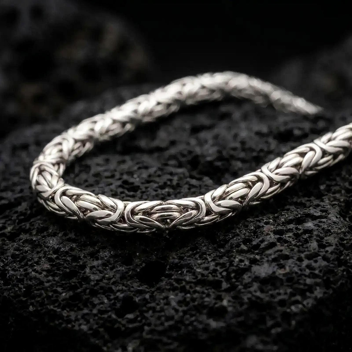 The polished silver 6mm Byzantine chain necklace is displayed across textured black volcanic rock, highlighting its dimensional round links and intricate woven construction in a premium macro photograph.