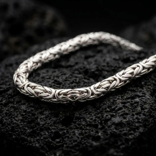 The polished silver 6mm Byzantine chain necklace is displayed across textured black volcanic rock, highlighting its dimensional round links and intricate woven construction in a premium macro photograph.