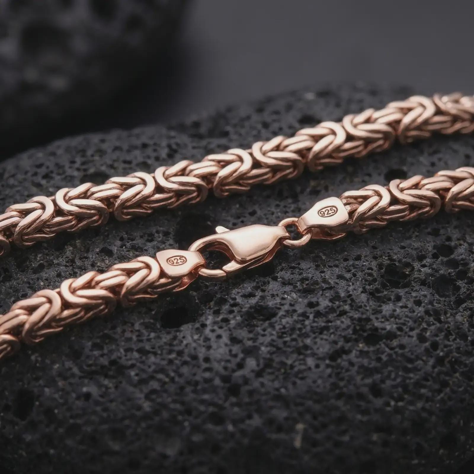 The rose gold plated 6mm Byzantine necklace is arranged on black volcanic rock, emphasizing its dimensional woven structure, polished clasp detail, and refined heavy-link construction in a macro image.