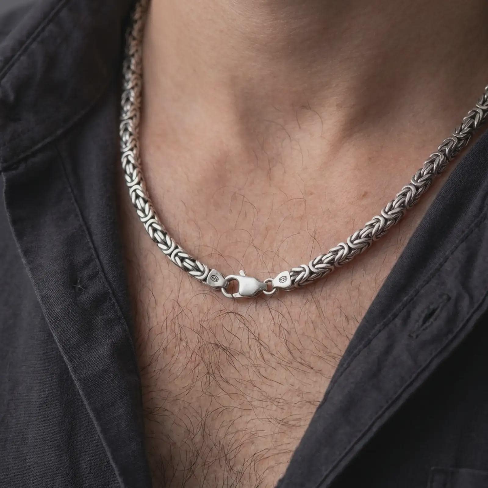 A 6mm Byzantine chain necklace with an oxidized finish is worn on a man’s neck, displaying dark recessed link details and a bold masculine silhouette in a close-up lifestyle shot.