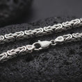 Load image into Gallery viewer, A 6mm Byzantine chain necklace with an oxidized finish rests on dark volcanic rock, showing the interwoven sterling silver links, 925 stamp, and lobster clasp in a macro studio close-up.
