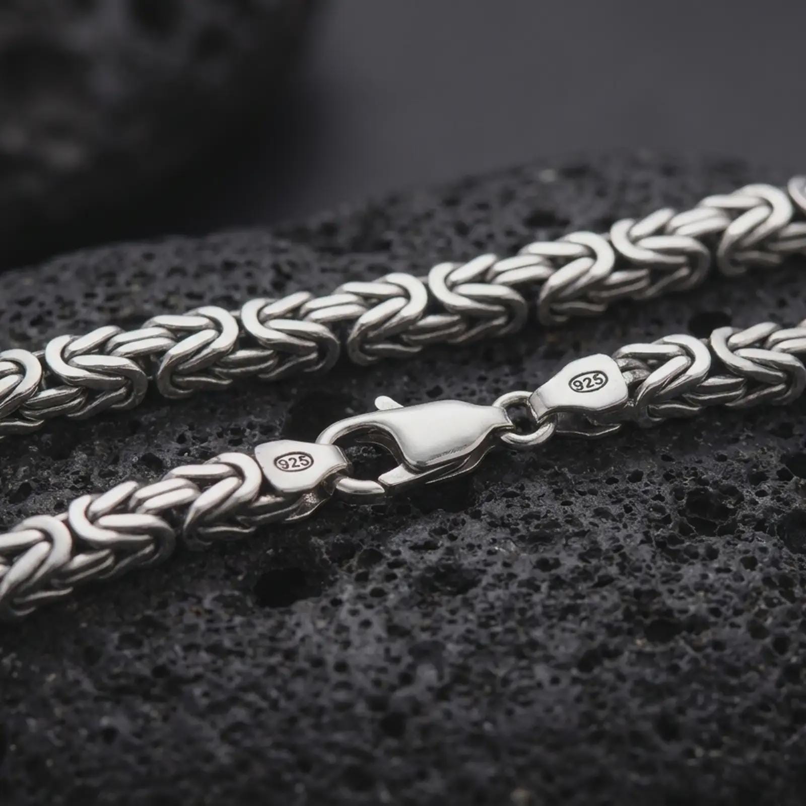 A 6mm Byzantine chain necklace with an oxidized finish rests on dark volcanic rock, showing the interwoven sterling silver links, 925 stamp, and lobster clasp in a macro studio close-up.