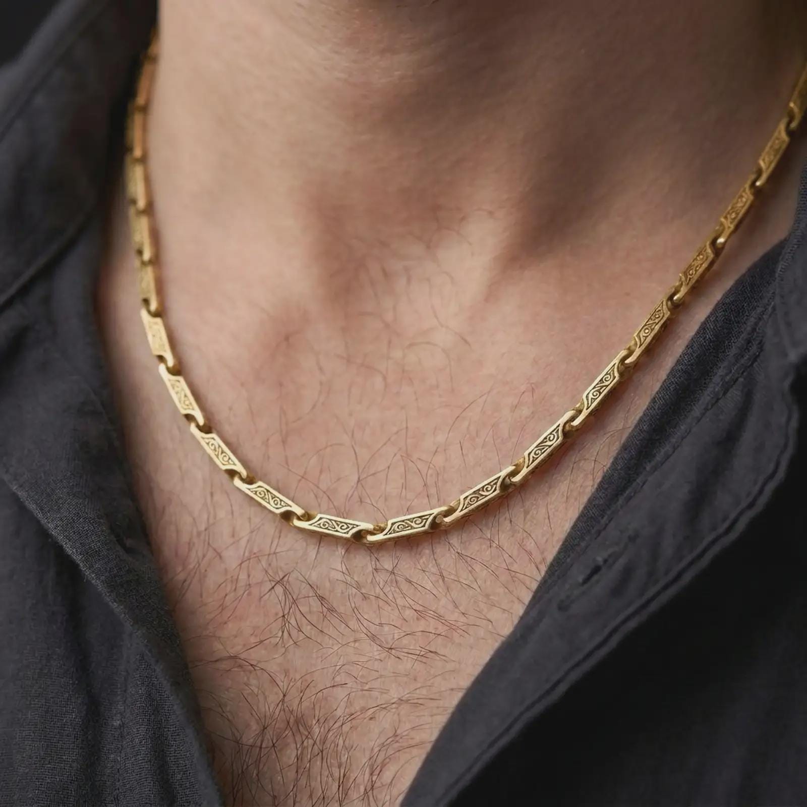 A gold plated scroll link chain necklace is worn on a man’s neck with dark clothing, highlighting the decorative bar links and strong statement jewelry style in a refined close-up.