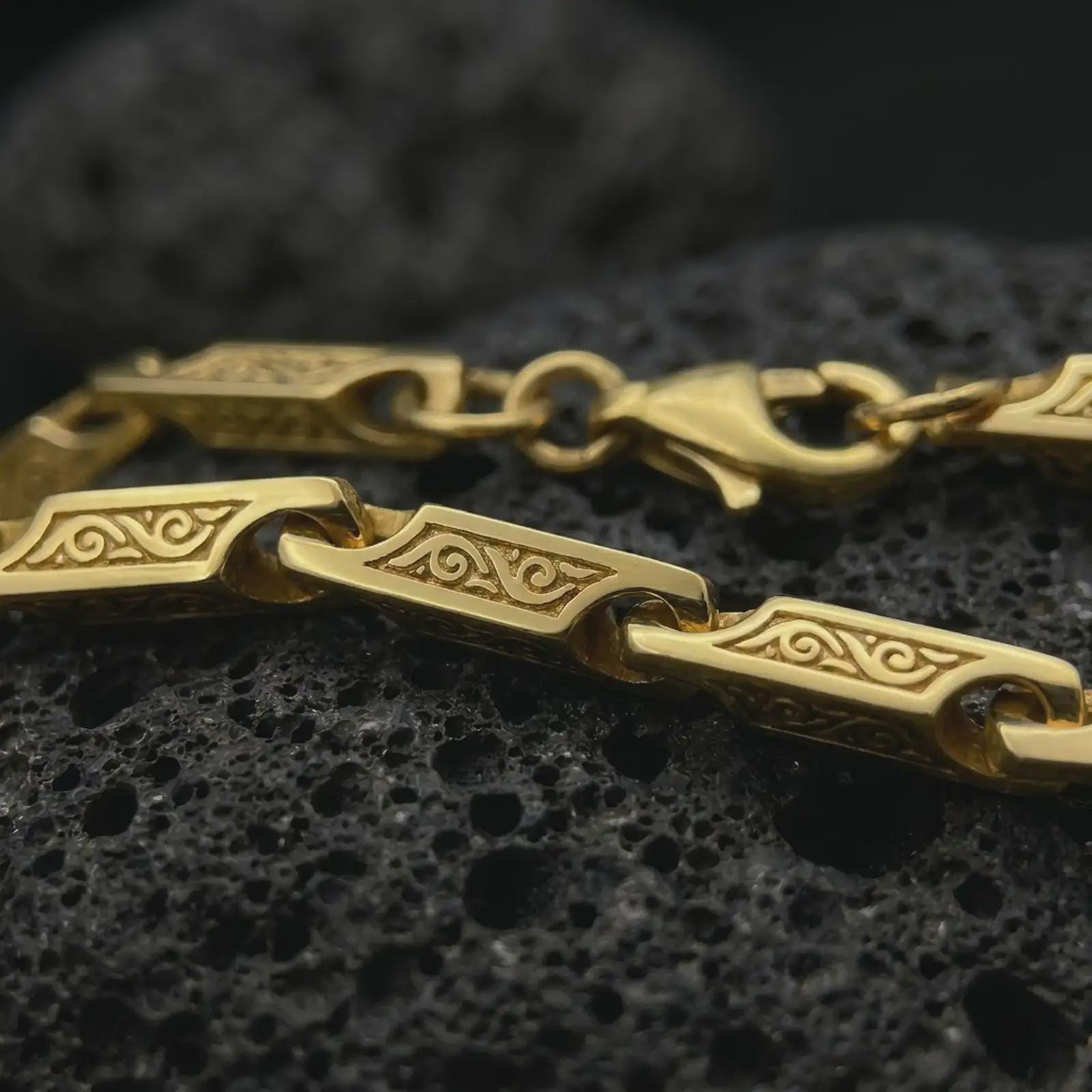 A gold plated ornate chain necklace lies across rugged volcanic rock, featuring rectangular decorative links with recessed scrollwork and a bold masculine profile in a luxury macro studio close-up.