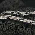 Load image into Gallery viewer, The polished silver bar link chain necklace is displayed on textured black volcanic rock, highlighting the flowing scroll engravings, smooth link openings, and structured sterling silver construction in a premium macro photograph.
