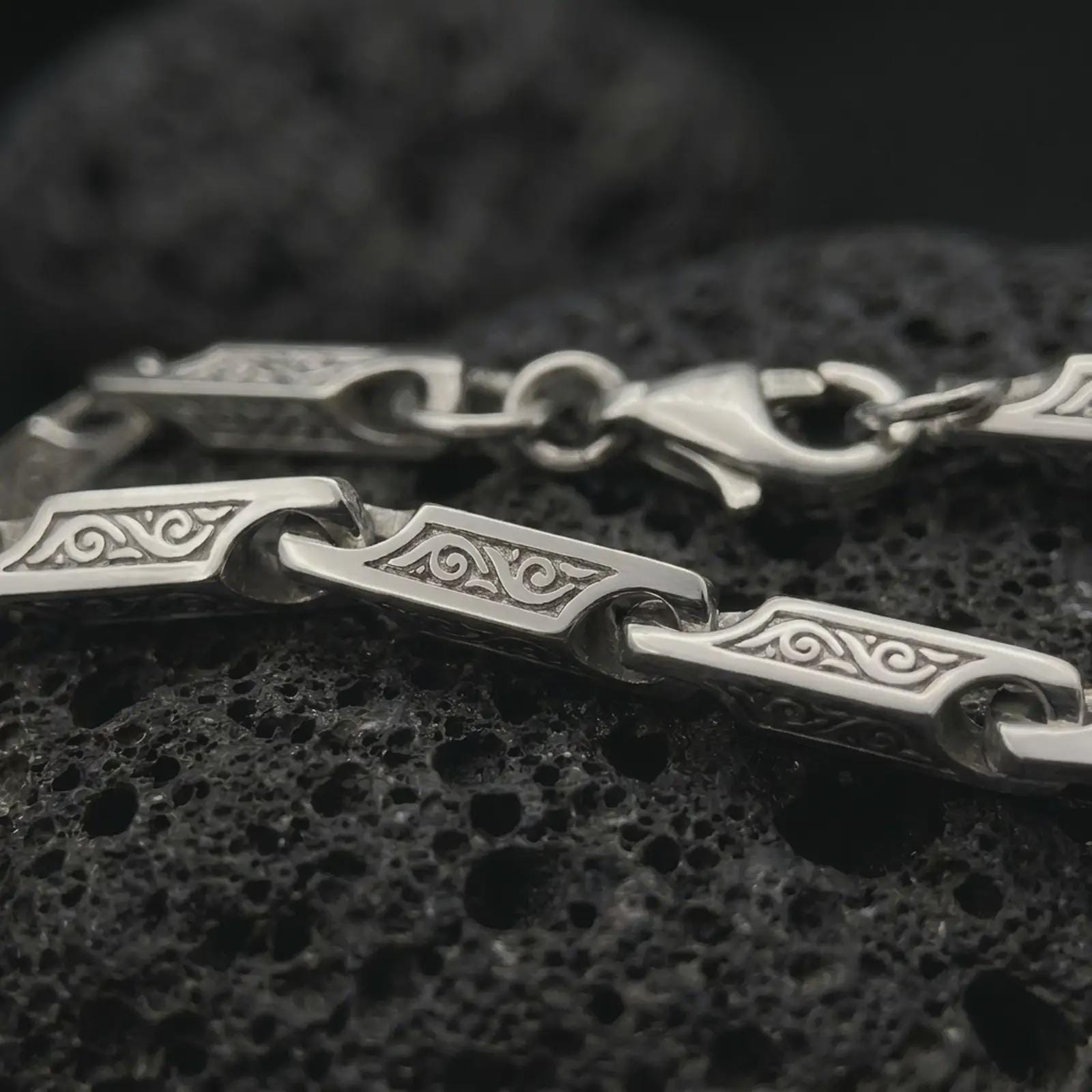 The polished silver bar link chain necklace is displayed on textured black volcanic rock, highlighting the flowing scroll engravings, smooth link openings, and structured sterling silver construction in a premium macro photograph.