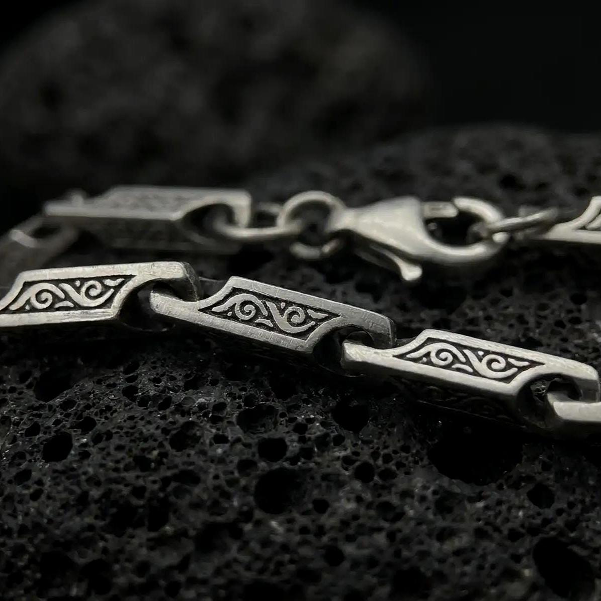 An oxidized ornate scroll chain necklace rests on dark volcanic rock, showing rectangular engraved bar links, recessed swirl details, and articulated connectors in a macro studio close-up.