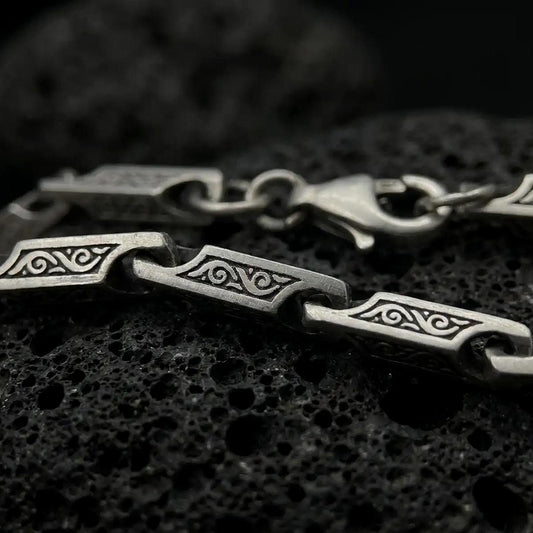 An oxidized ornate scroll chain necklace rests on dark volcanic rock, showing rectangular engraved bar links, recessed swirl details, and articulated connectors in a macro studio close-up.