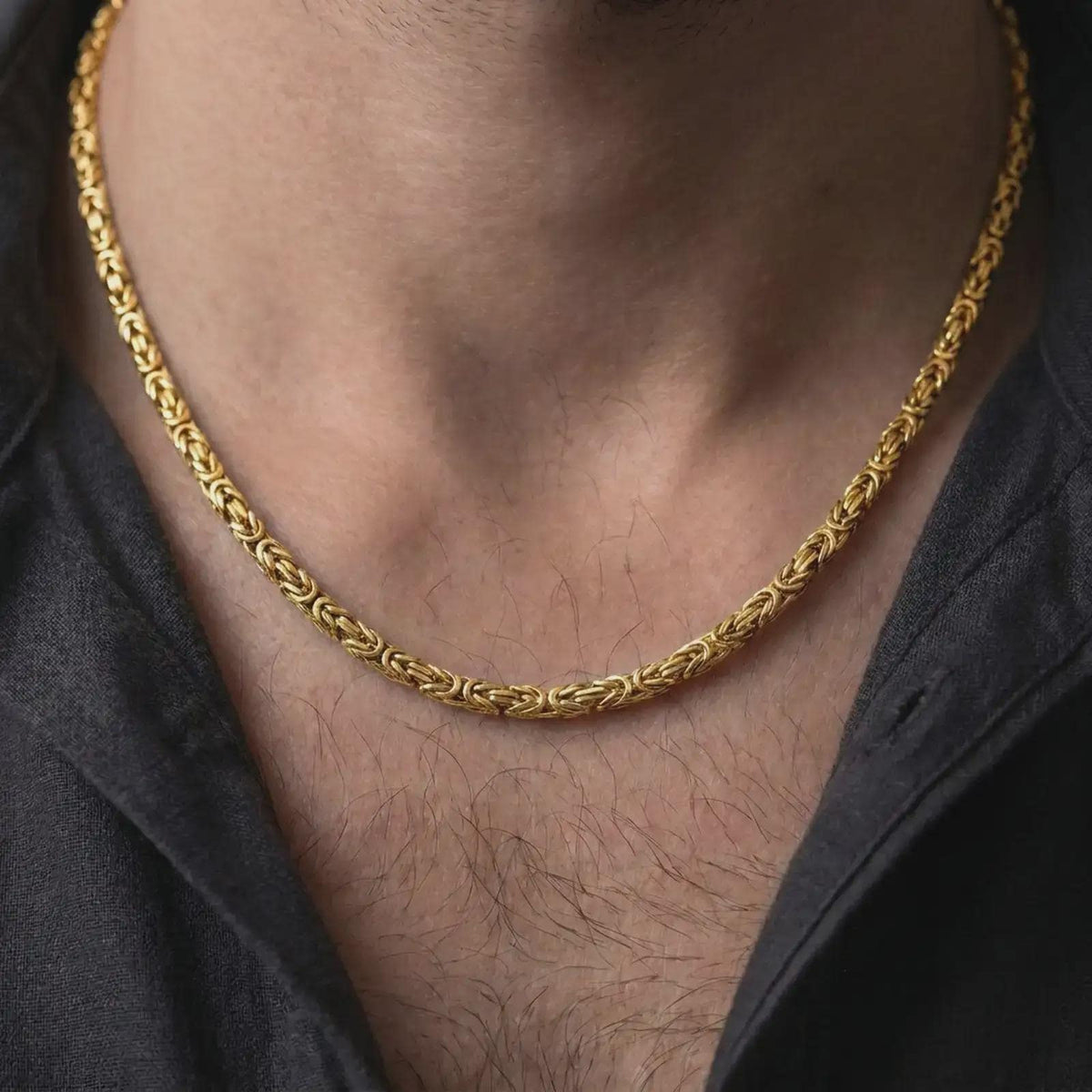 A gold plated 4mm square Byzantine chain necklace is worn on a man with dark clothing, emphasizing its woven link texture and bold statement appeal in a close-up lifestyle image.