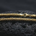 Load image into Gallery viewer, A gold plated 4mm square Byzantine chain necklace lies across rugged volcanic rock, showcasing its interwoven link design and strong masculine profile in a luxury macro close-up.
